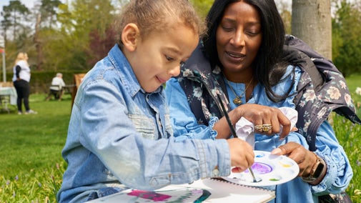 A woman and child sit outdoors on a grassy field with trees behind them, painting a colourful picture